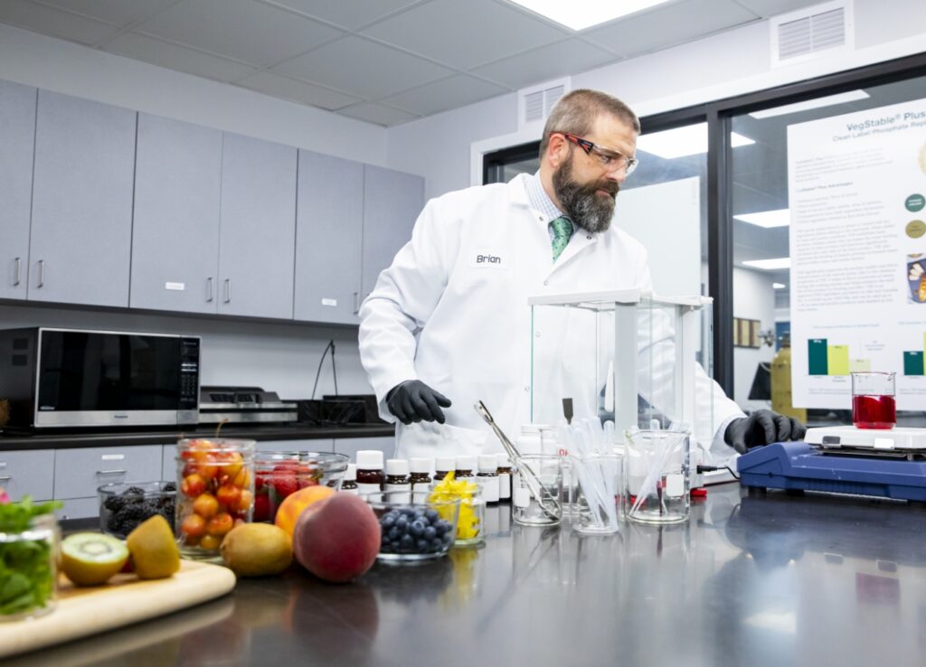 man in a lab coat standing in front of a counter filled with fruits and vegetables, showcasing fresh ingredients for beverages and manufacturing