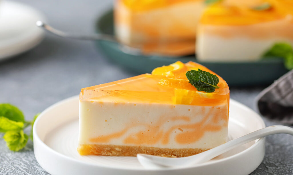 Mango cheesecake slice with orange gelatin swirl topping, mint, and citrus garnish on a white plate.