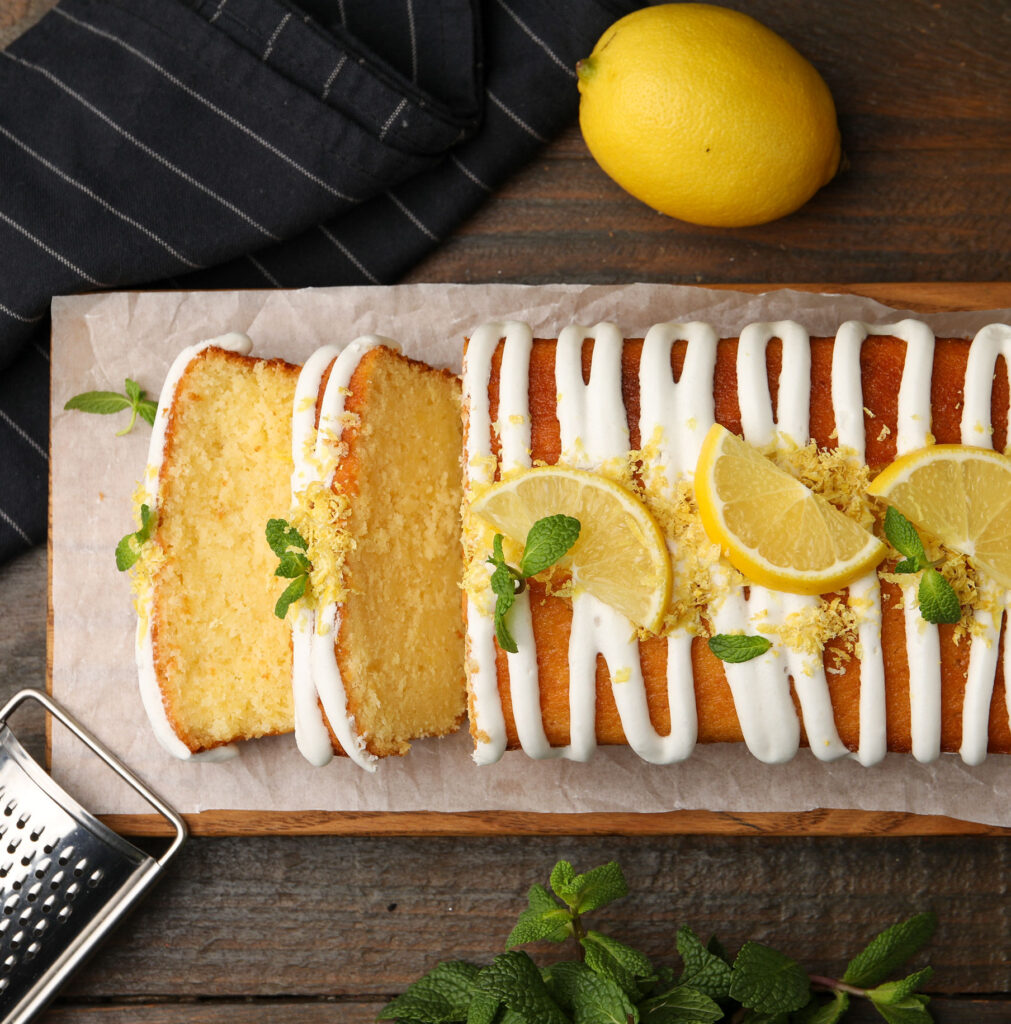 Sliced lemon zest pound cake with white glaze, topped with fresh lemon slices and mint on a wood board.