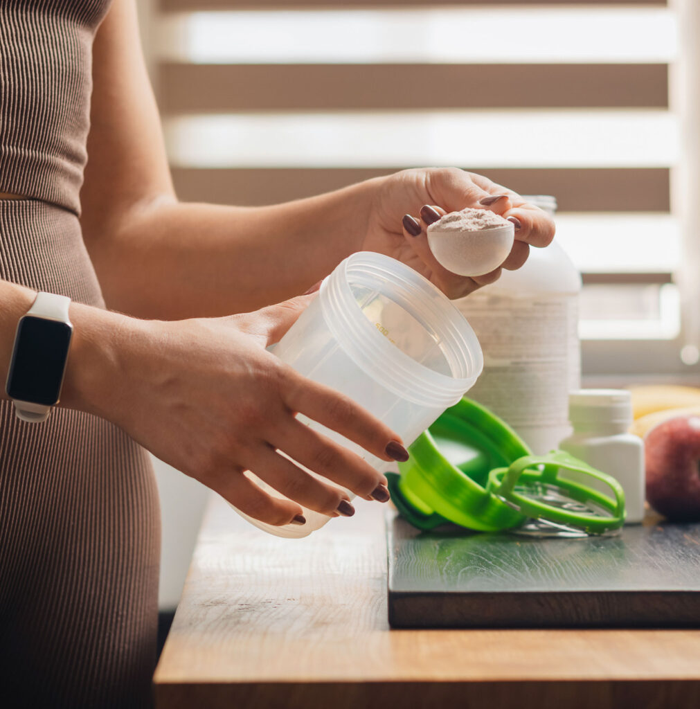 Person scooping protein powder into a shaker bottle with supplements and fruit on a table.